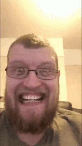 A Man With A Beard And Glasses Is Smiling And Looking At The Camera . GIF
