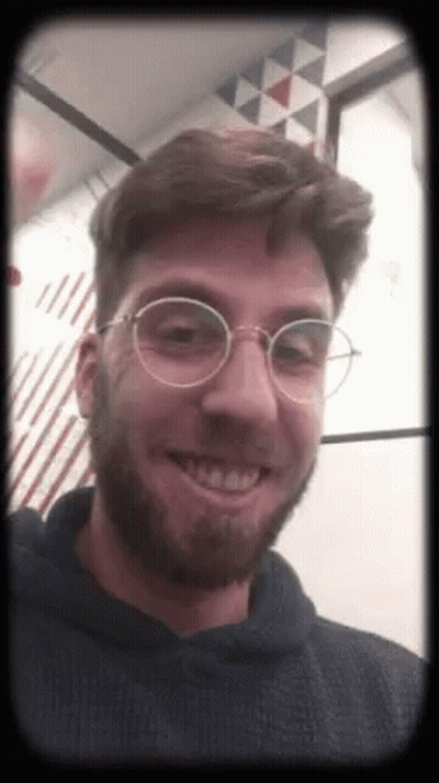A Man With A Beard And Glasses Is Smiling And Wearing A Black Hoodie . GIF