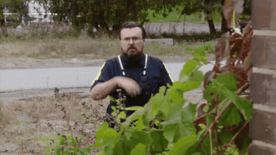 A Man With A Beard And Glasses Is Standing In Front Of A Plant . GIF