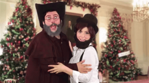 A Man With A Beard And Hat Is Hugging A Woman In Front Of Christmas Trees GIF