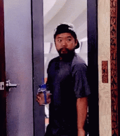A Man With A Beard And Hat Is Standing In A Doorway Holding A Bottle Of Water . GIF