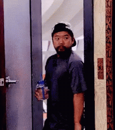 A Man With A Beard And Hat Is Standing In A Doorway Holding A Bottle Of Water . GIF