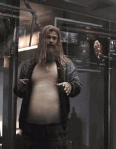 A Man With A Beard And Long Hair Is Standing In A Dark Room And Pointing At His Belly . GIF