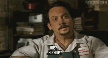 A Man With A Beard And Mustache Is Sitting At A Table Holding A Book And Talking About Hip Hop . GIF