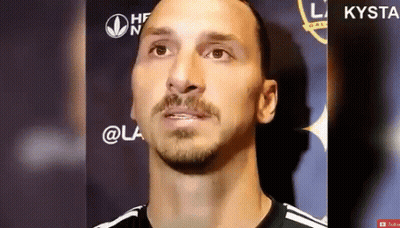 A Man With A Beard And Mustache Is Standing In Front Of A Sign That Says Kysta GIF