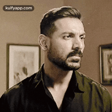 A Man With A Beard And Mustache Is Standing In Front Of A Wall With Pictures On It . GIF