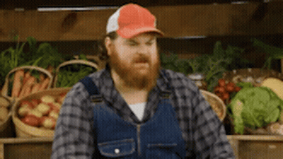 A Man With A Beard And Overalls Is Standing In Front Of A Fruit Stand . GIF