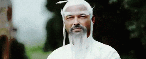 A Man With A Beard And White Hair Is Wearing A White Robe And A White Hat . GIF