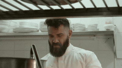 A Man With A Beard In A Kitchen With Plates On The Shelf Behind Him GIF