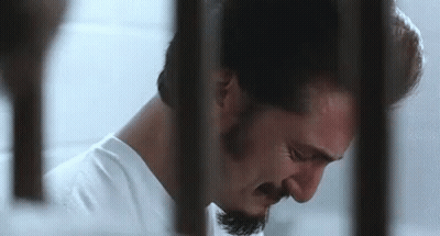A Man With A Beard Is Behind Bars In A Jail Cell . GIF