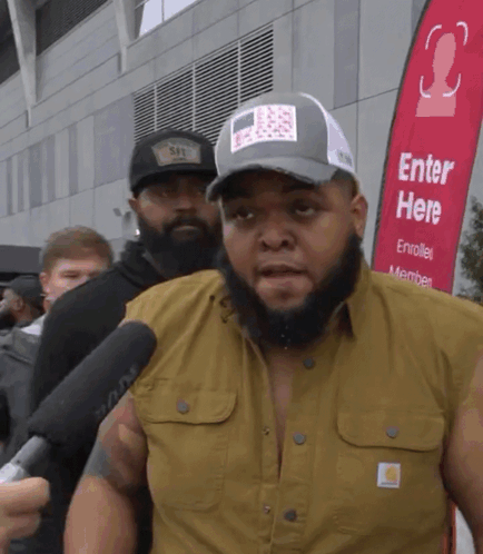 A Man With A Beard Is Being Interviewed In Front Of A Red Banner That Says Enter Here GIF