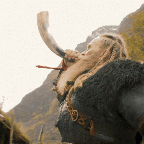A Man With A Beard Is Blowing A Horn In Front Of Mountains GIF