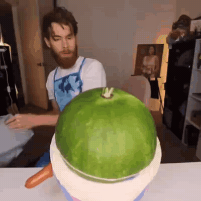 A Man With A Beard Is Carving A Watermelon With A Carrot On Top Of It . GIF