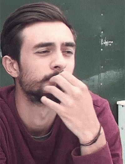A Man With A Beard Is Covering His Mouth With His Hand While Wearing A Maroon Shirt . GIF