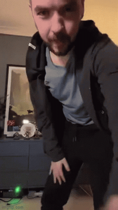 A Man With A Beard Is Dancing In A Living Room While Wearing A Black Jacket And A Blue Shirt . GIF