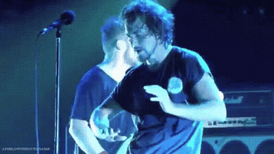 A Man With A Beard Is Dancing On A Stage In Front Of A Microphone While Another Man Stands Behind Him . GIF