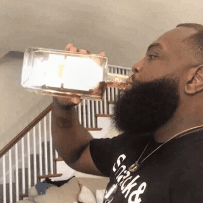 A Man With A Beard Is Drinking From A Bottle In Front Of A Staircase . GIF