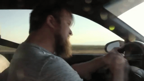 A Man With A Beard Is Driving A Car And Looking Out The Window GIF
