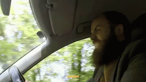 A Man With A Beard Is Driving A Car With His Eyes Closed And A Sticker That Says Noosh GIF