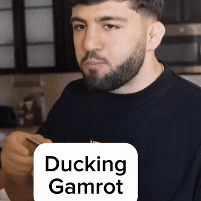 A Man With A Beard Is Holding A Cell Phone In Front Of A Sign That Reads Ducking Gamrot GIF