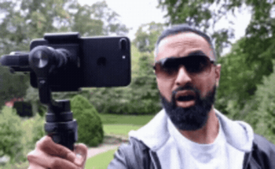 A Man With A Beard Is Holding A Cell Phone On A Tripod While Wearing Sunglasses . GIF