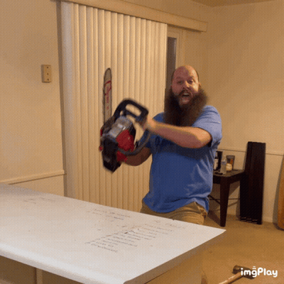 A Man With A Beard Is Holding A Chainsaw In Front Of A Window GIF