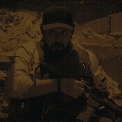 A Man With A Beard Is Holding A Gun And Wearing A Hat With The Letter T On It GIF