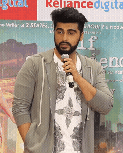 A Man With A Beard Is Holding A Microphone In Front Of A Sign That Says Reliance Digital GIF