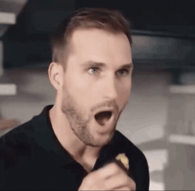 A Man With A Beard Is Holding A Piece Of Food In His Hand And Making A Surprised Face . GIF