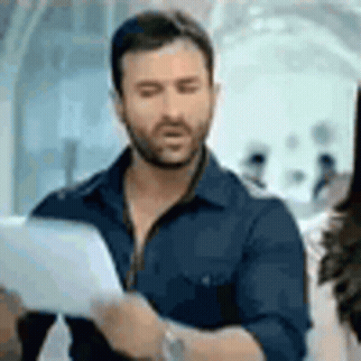A Man With A Beard Is Holding A Piece Of Paper And Looking At It . GIF