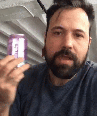 A Man With A Beard Is Holding A Purple Can Of Soda With The Letter T On It GIF