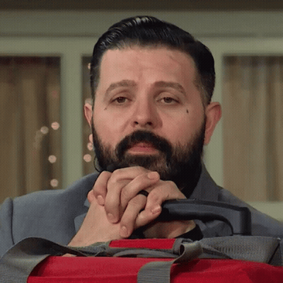 A Man With A Beard Is Holding A Red Suitcase With His Hands Folded . GIF