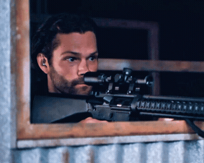 A Man With A Beard Is Holding A Rifle In Front Of A Window GIF