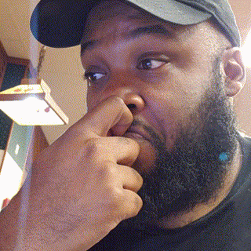A Man With A Beard Is Holding His Nose While Wearing A Baseball Cap GIF