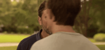 A Man With A Beard Is Kissing Another Man On The Forehead In A Park . GIF