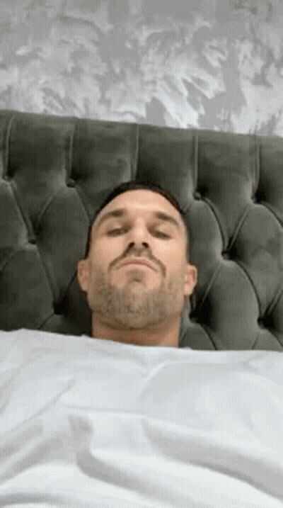 A Man With A Beard Is Laying In Bed With His Eyes Closed And His Head On The Headboard . GIF