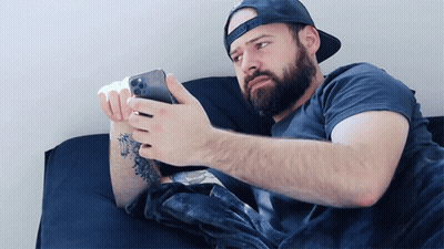 A Man With A Beard Is Laying On A Bed Looking At A Cell Phone GIF