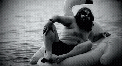 A Man With A Beard Is Laying On An Inflatable Swan In The Water . GIF