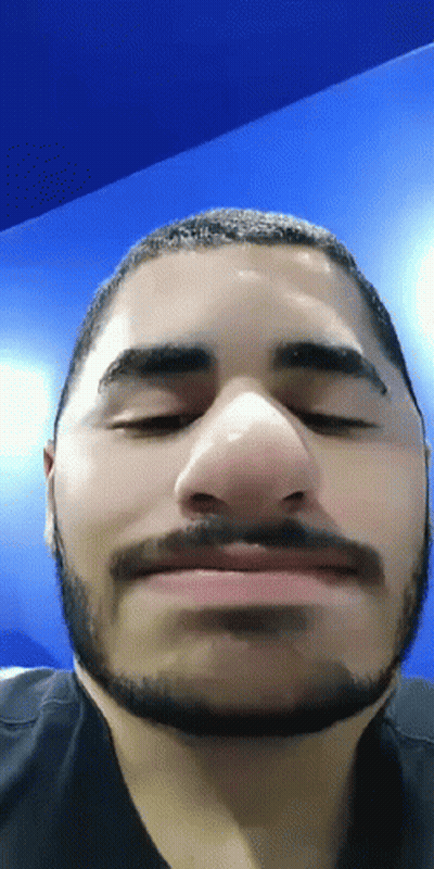 A Man With A Beard Is Making A Funny Face With His Eyes Closed In Front Of A Blue Wall . GIF