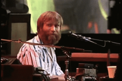 A Man With A Beard Is Playing A Keyboard And Singing Into A Microphone On A Stage . GIF