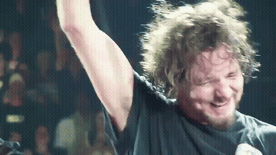 A Man With A Beard Is Singing Into A Microphone With His Arm Up In The Air GIF