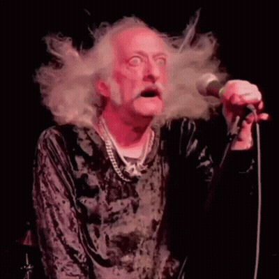 A Man With A Beard Is Singing Into A Microphone With His Hair Blowing In The Wind . GIF
