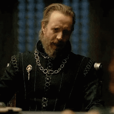 A Man With A Beard Is Sitting At A Table Wearing A Black Jacket And A Gold Chain Around His Neck . GIF