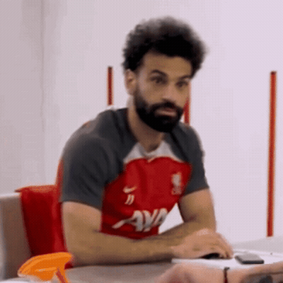 A Man With A Beard Is Sitting At A Table Wearing A Red Shirt That Says Ava On It GIF