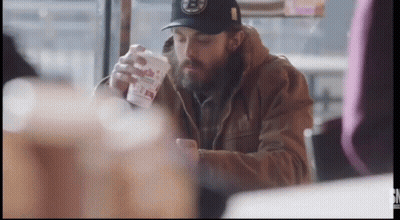 A Man With A Beard Is Sitting At A Table With A Cup In His Hand . GIF