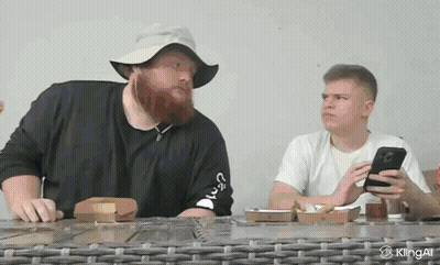 A Man With A Beard Is Sitting At A Table With A Phone In His Hand GIF