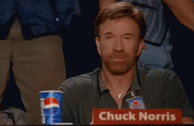 A Man With A Beard Is Sitting At A Table With A Sign That Says Chuck Norris GIF
