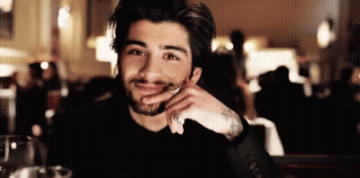 A Man With A Beard Is Sitting At A Table With His Hand On His Chin And Smiling At The Camera . GIF