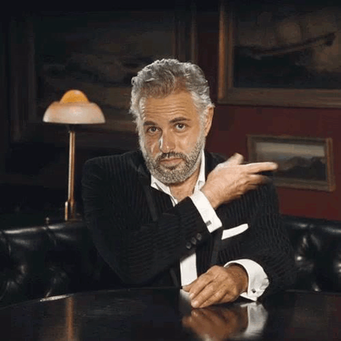 A Man With A Beard Is Sitting At A Table With His Hands In His Pockets And A Lamp In The Background . GIF