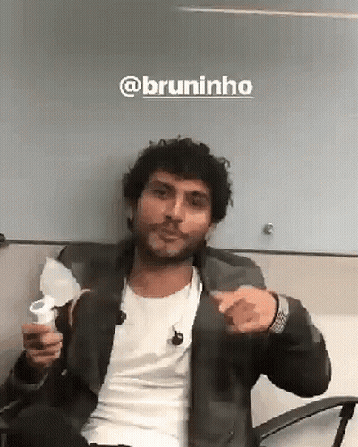A Man With A Beard Is Sitting In A Chair Holding A Cup And Pointing . GIF